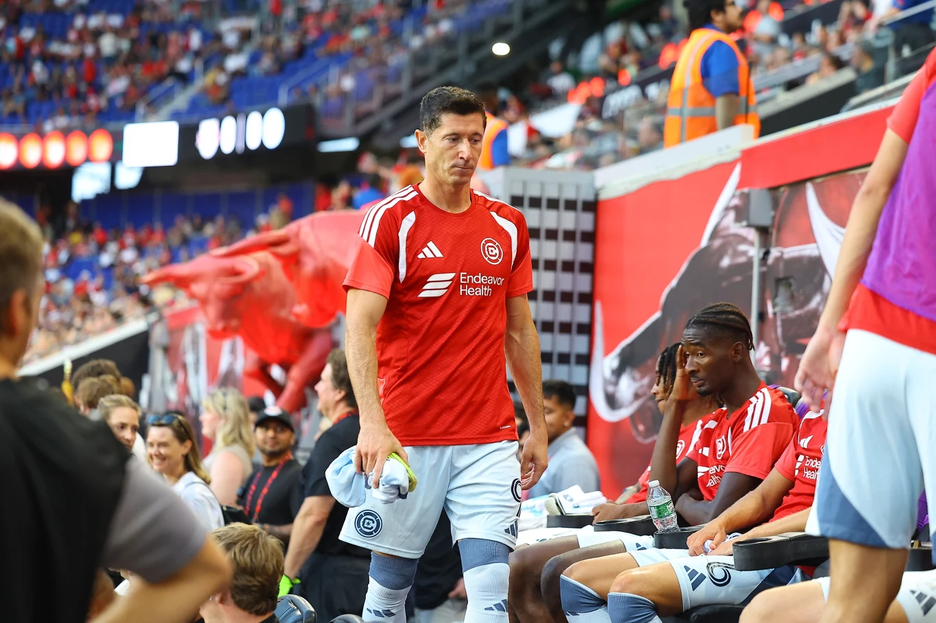 Robert Lewandowski w Chicago Fire Piłkarz w czerwonej koszulce z logo klubowym opuszcza ławkę rezerwowych, podczas gdy pozostali zawodnicy, również w strojach drużyny, siedzą obok na ławce; w tle trybuny z kibicami i stadionowe dekoracje.