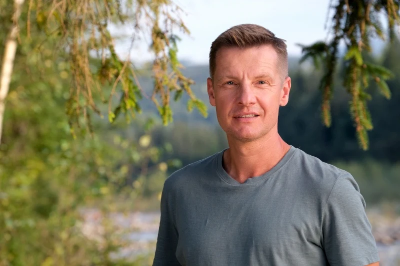 Piotr nie pozwoli Kindze czekać z wizytą u lekarza Uśmiechnięty mężczyzna w szarej koszulce stoi wśród zieleni, otoczony gałęziami drzew, z rozmytym tłem natury za sobą.