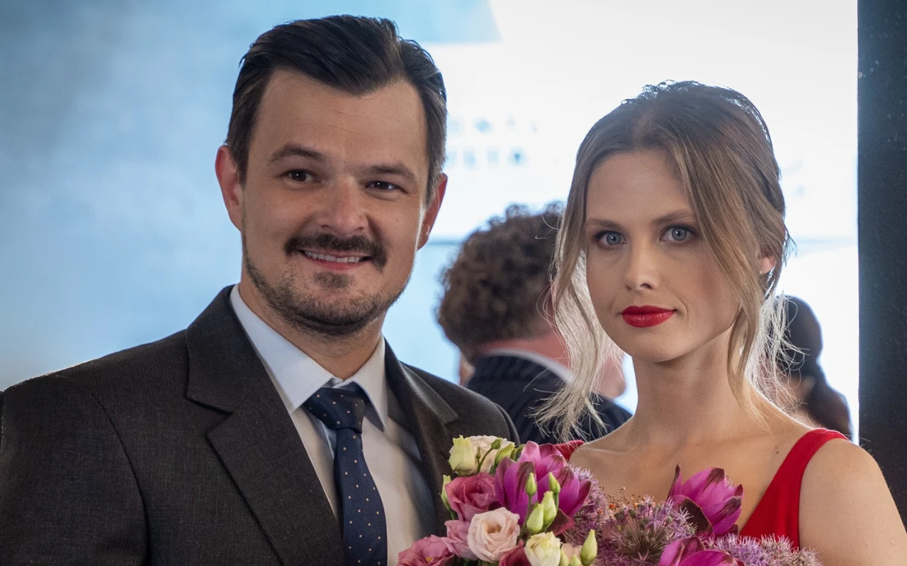 Uśmiechnięty mężczyzna w garniturze i kobieta w eleganckiej czerwonej sukni z bukietem kolorowych kwiatów w rękach, oboje zwróceni w stronę obiektywu.
