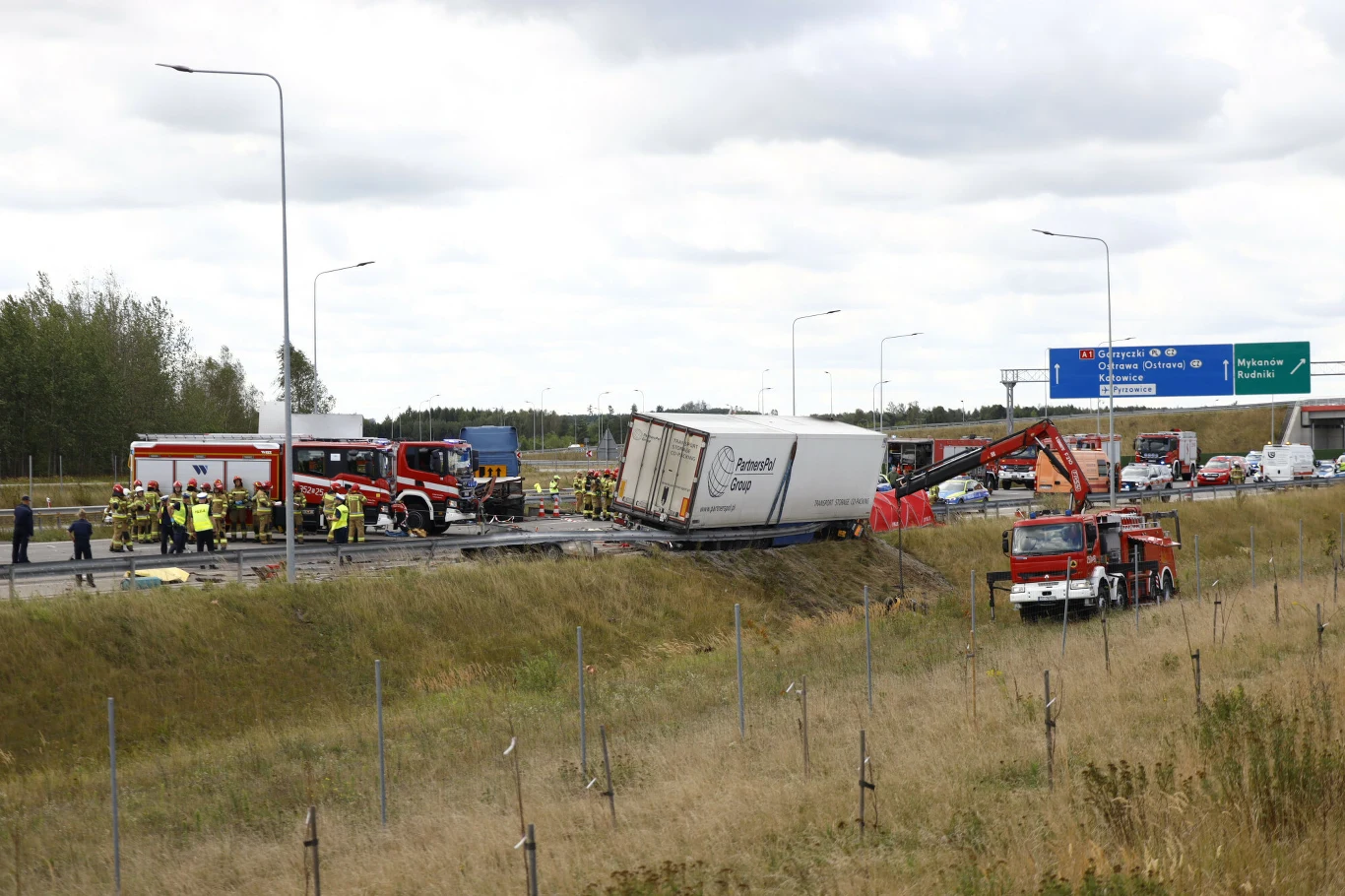 Służby ratownicze pracujące przy ciężarówce leżącej na boku na autostradzie, wspierane przez dźwigi i wozy strażackie, w otoczeniu zablokowanego pasa ruchu.