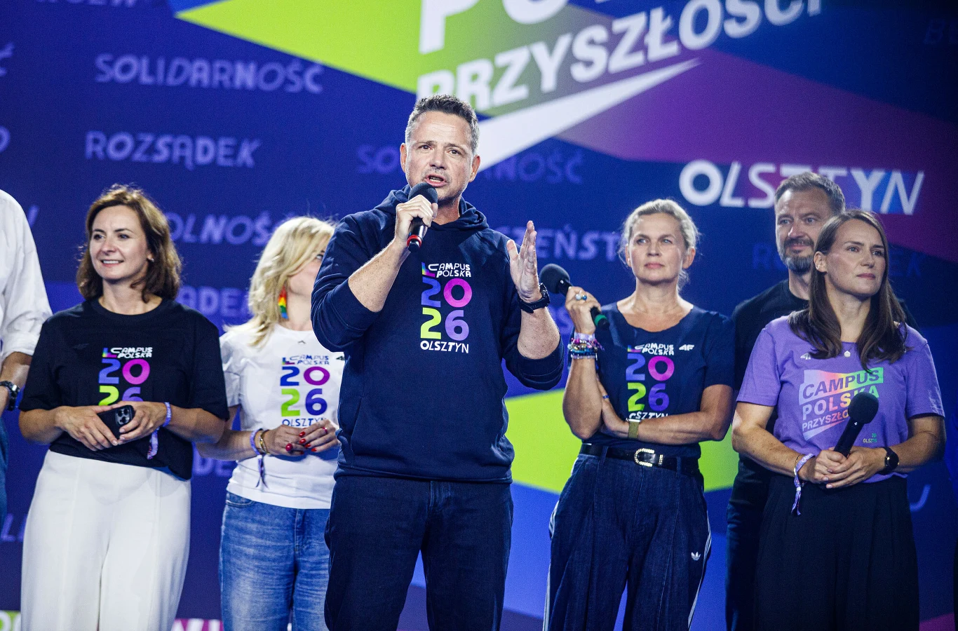 Rafał Trzaskowski podczas Campusu Polska Rafał Trzaskowski przemawia na scenie podczas Campusu Polska Przyszłości w Olsztynie, obok grupa osób w koszulkach z logo.