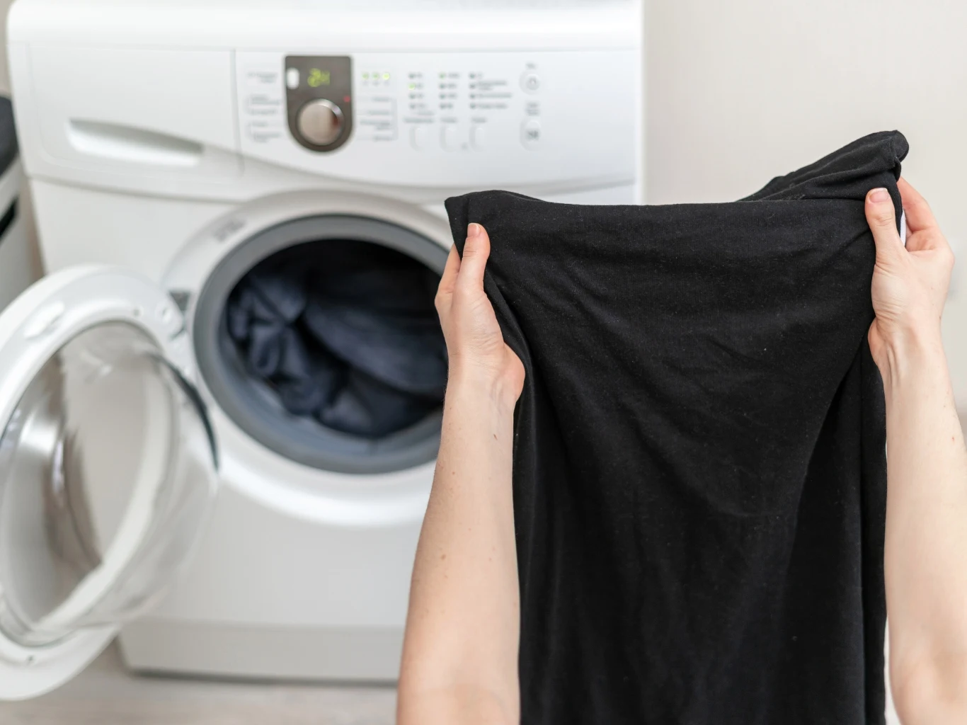Czarne ubrania najlepiej prać w niskiej temperaturze. Otwarte drzwiczki pralki, ręce wkładające czarną koszulkę do bębna z widoczną ciemną odzieżą.