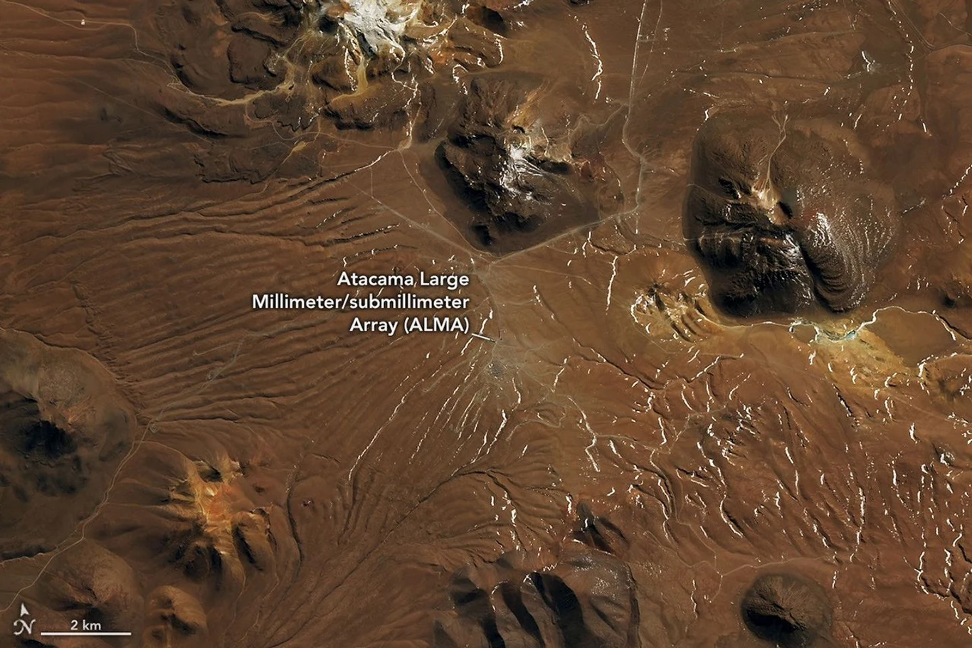 Obserwatorium Atacama Large Millimeter/submillimeter Array (ALMA) położone na pustynnym, górzystym terenie z licznymi jasnymi liniami wyżłobionymi w ziemi i wyraźnie widocznymi wierzchołkami okolicznych wzgórz.