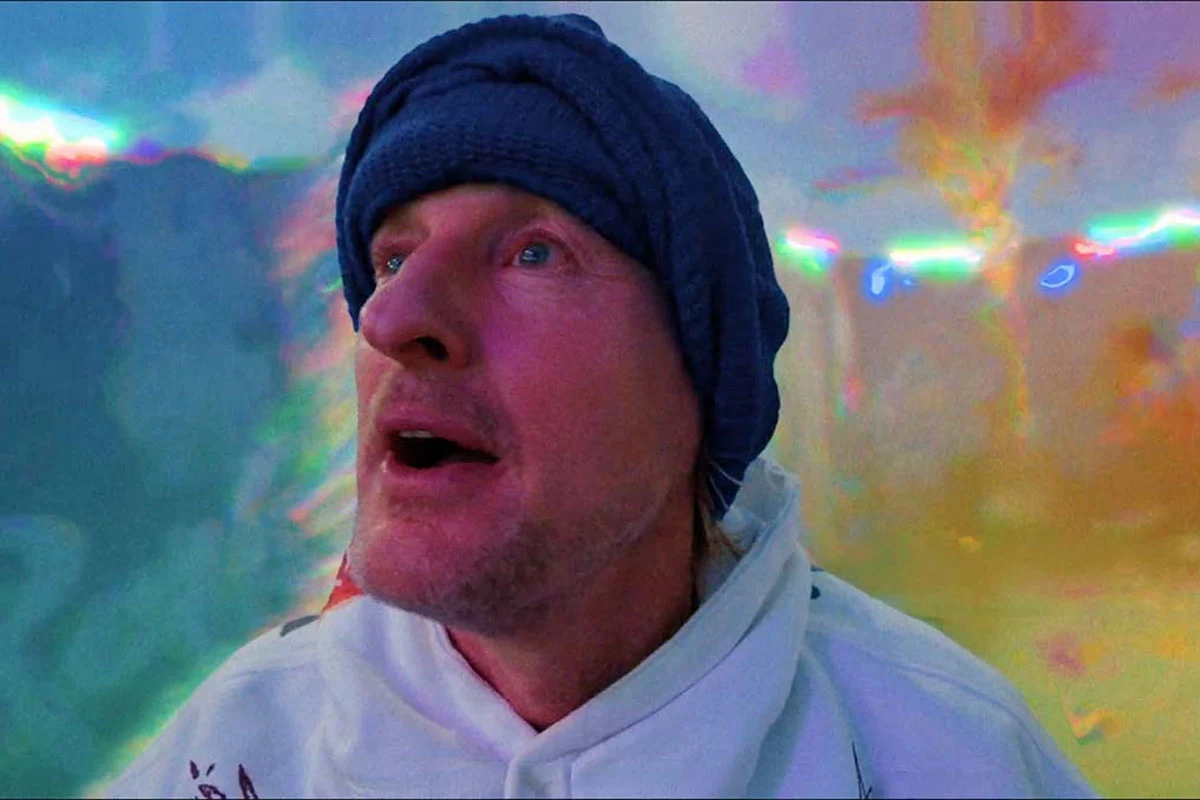 Owen Wilson w scenie z "Rolling Loud. Film" Owen Wilson w białej bluzie i granatowej czapce na tle kolorowego, rozmytego światła w scenie z filmu „Rolling Loud. Film”.