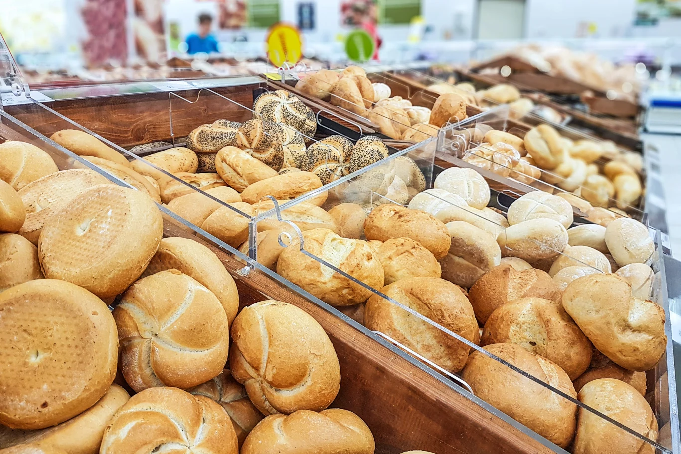 Rzędy różnorodnych bułek i pieczywa ułożone w przezroczystych pojemnikach w sklepowej piekarni.