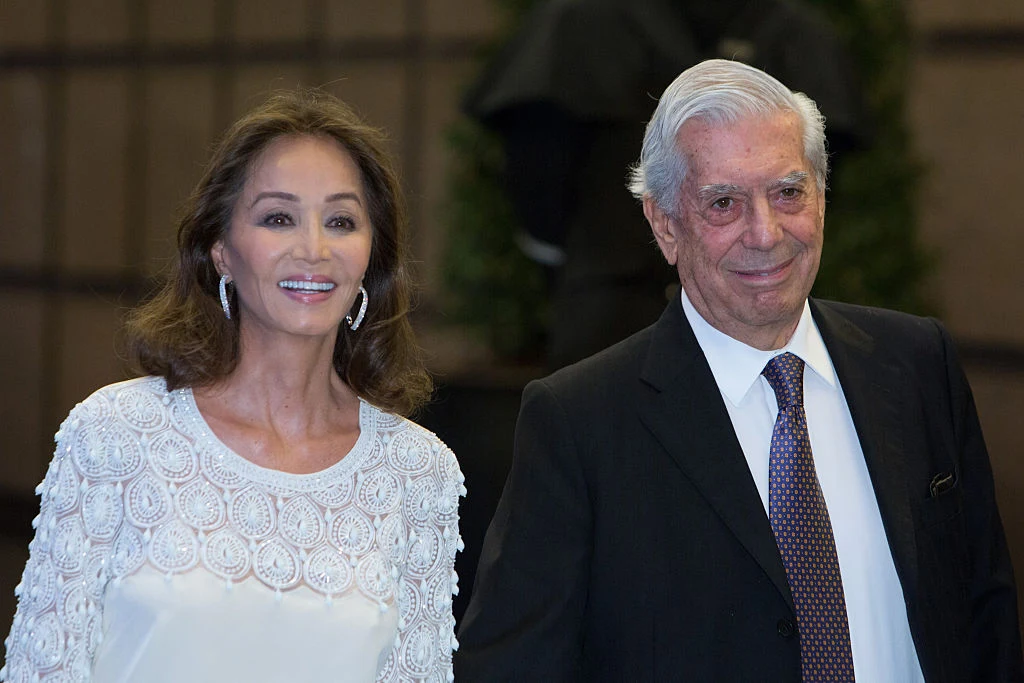 Mario Vargas Llosa i jego partnerka Isabel Preysler w 2016 roku Elegancka kobieta w białej, ozdobnej sukni i mężczyzna w ciemnym garniturze z krawatem stoją obok siebie i uśmiechają się w kierunku aparatu.