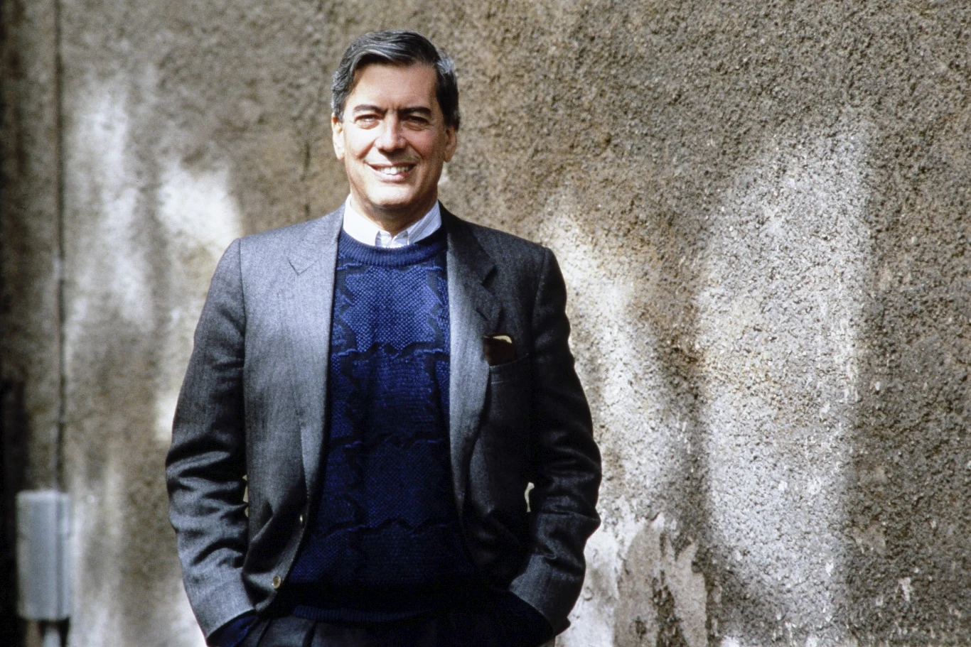 Mario Vargas Llosa w 1987 roku Uśmiechnięty mężczyzna w marynarce i swetrze stoi oparty o szarą, betonową ścianę.