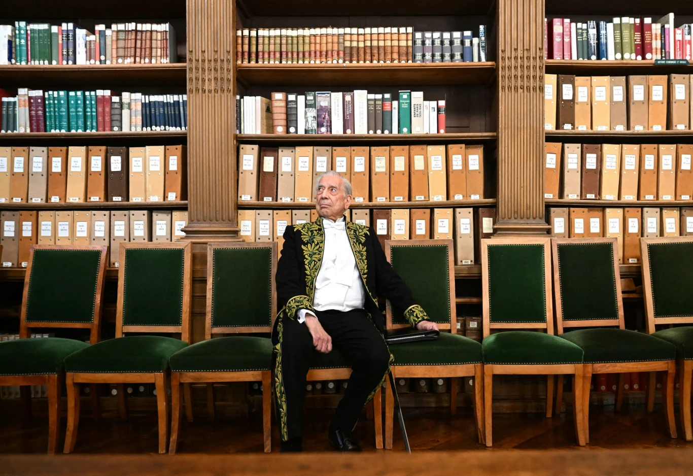 Mario Vargas Llosa Starszy mężczyzna w ciemnym stroju z ozdobnymi złotymi haftami siedzi samotnie na zielonym krześle, otoczony pustymi fotelami, na tle wysokiego regału wypełnionego książkami i segregatorami.