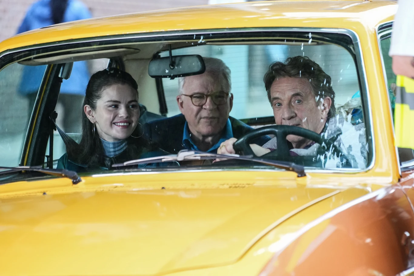 Selena Gomez, Steve Martin i Martin Short Trzy osoby siedzą wewnątrz żółtego samochodu, dwoje mężczyzn na przednich siedzeniach oraz uśmiechnięta kobieta obok