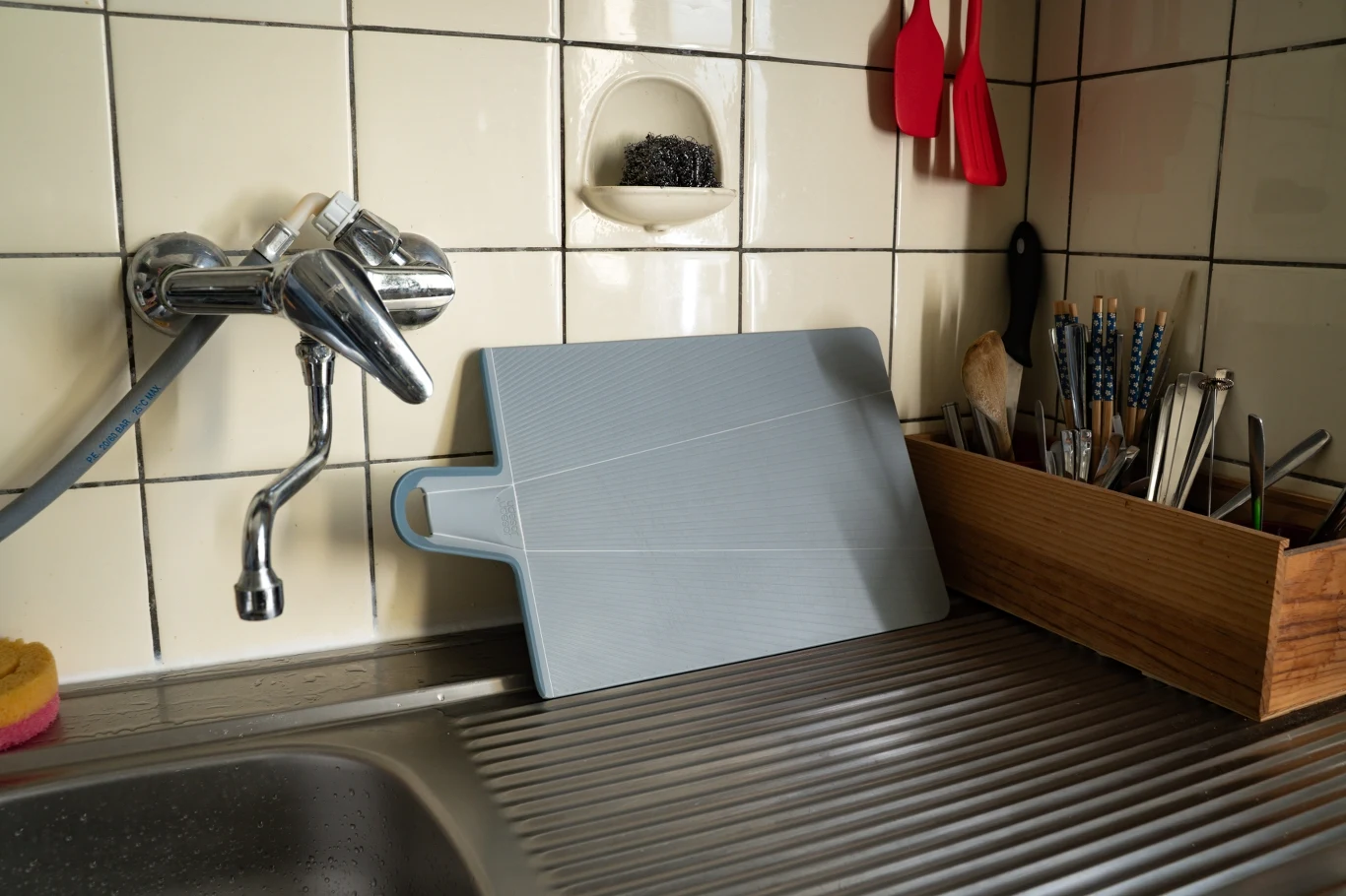 Szara deska do krojenia oparta o ścianę przy zlewie, obok drewniane pudełko z suszącymi się sztućcami i przyborami kuchennymi, na ścianie metalowa szczotka oraz zawieszone czerwone łopatki.