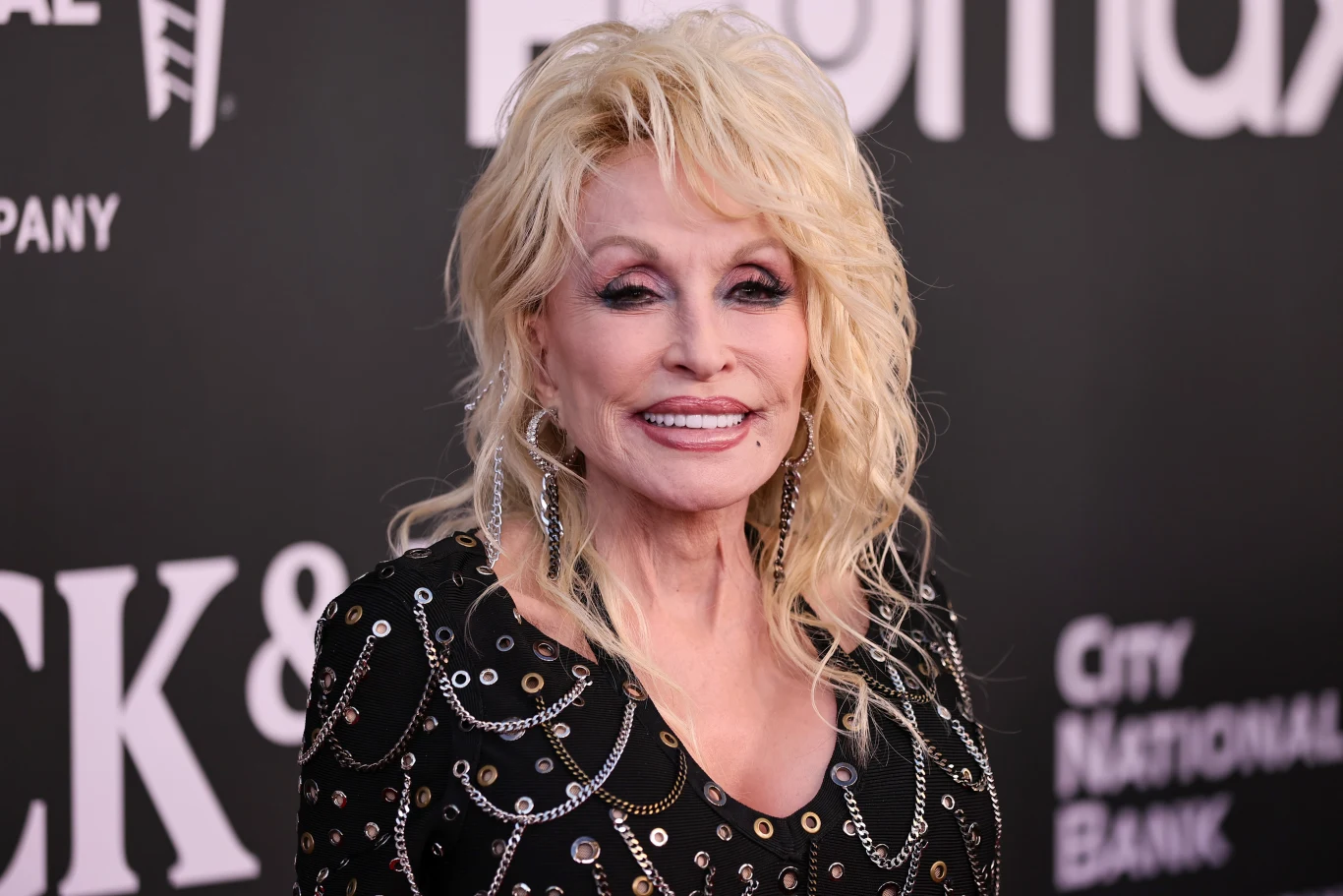 Dolly Parton zmarła w wieku 80 lat Dolly Parton, blond włosy, czarna suknia z błyszczącymi elementami, na tle ścianki z logotypami.