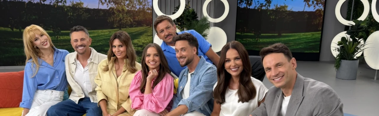 Specjalne wydanie "halo tu polsat". Wszystkie pary prowadzących razem Osiem uśmiechniętych osób siedzących razem na kolorowej kanapie we wnętrzu studia telewizyjnego, w tle ekrany z widokiem zielonego krajobrazu i nowoczesne dekoracje.