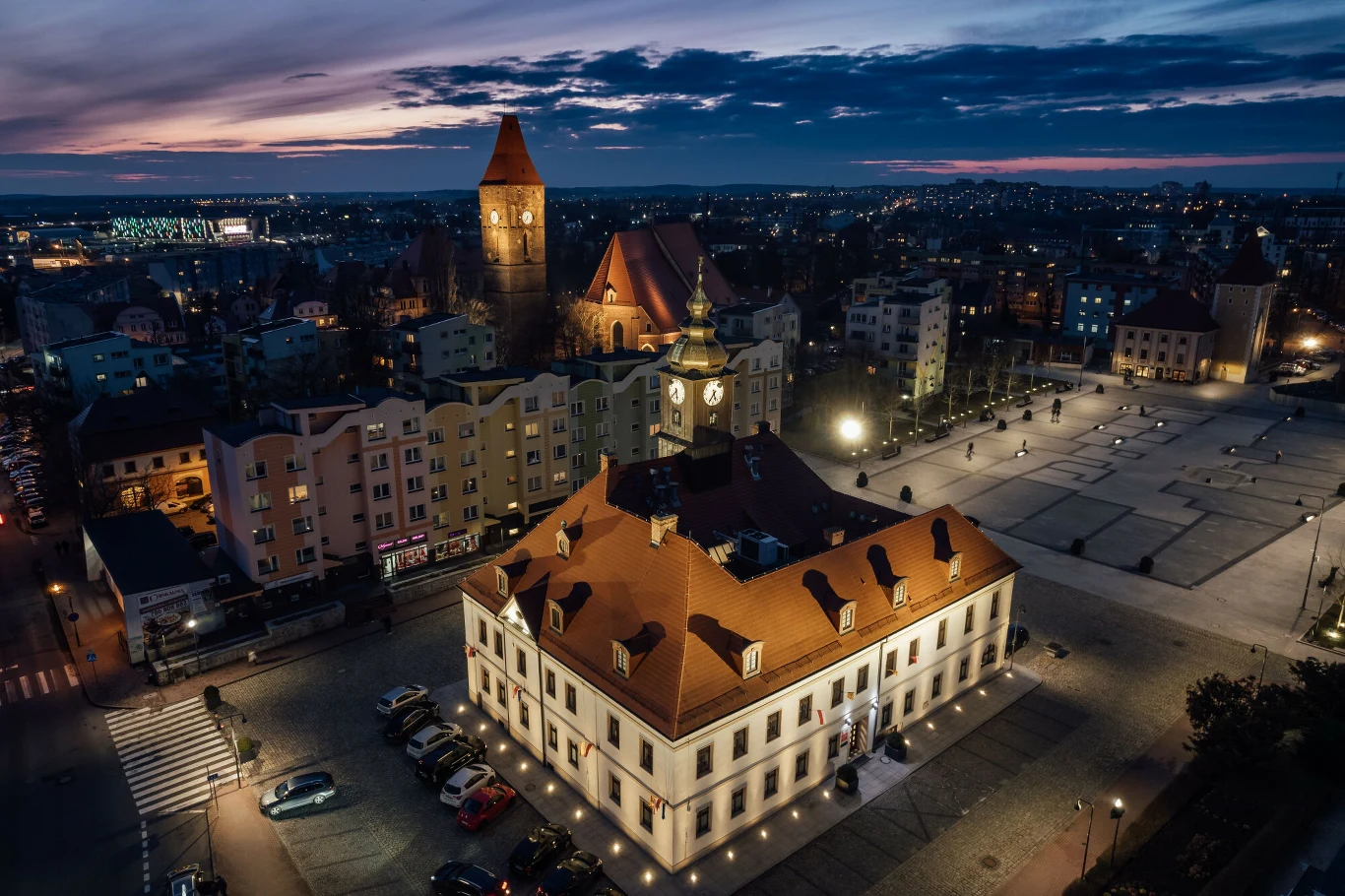 Rynek Lubina z lotu ptaka Oświetlony ratusz z zegarem i czerwonym dachem otoczony pustym placem miejskim, w tle kościół i zabudowa miasta.