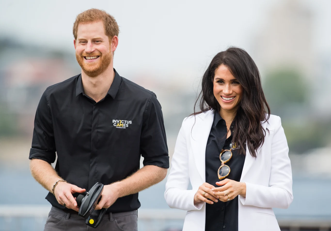 Start nowego życia w Wielkiej Brytanii nie jest łatwy dla Meghan i Harry'ego Uśmiechnięta para ubrana w casualowe, eleganckie stroje, mężczyzna w czarnej koszuli z logo Invictus Games i kobieta w białej marynarce, stoją obok siebie, prezentując swobodną i przyjazną atmosferę.