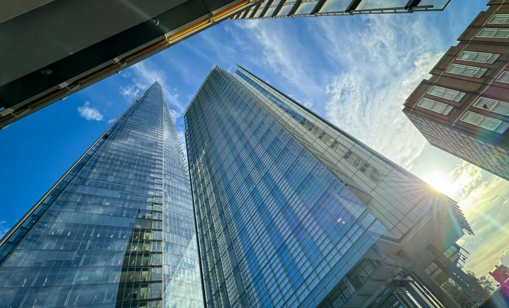 Apartamentowiec Shard Place w Londynie (pośrodku) Dwie wysokie, nowoczesne wieżowce ze szklaną elewacją odbijające niebo i chmury, u ich podnóża słońce przebijające się przez zabudowania po prawej stronie kadru.