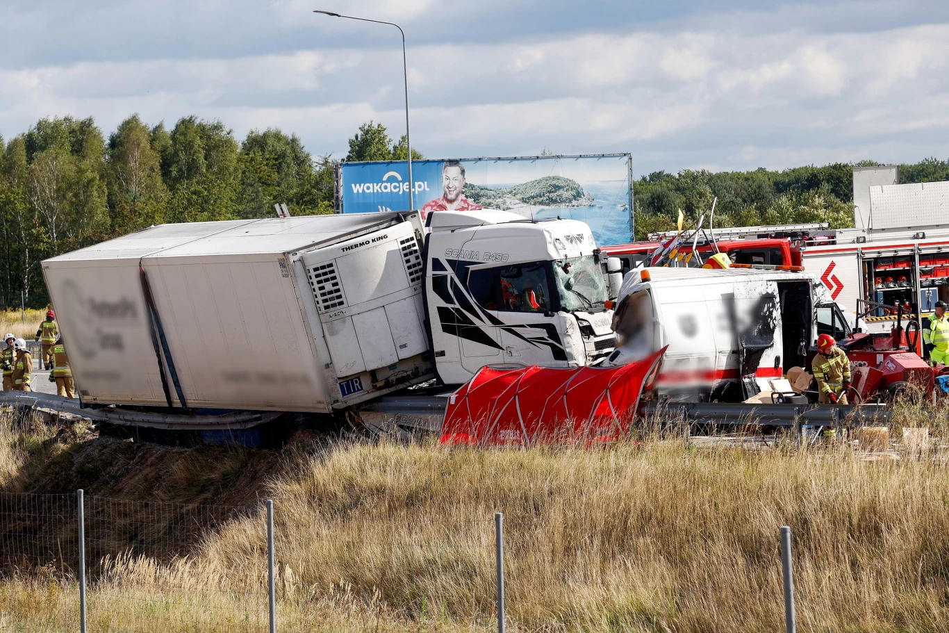 Ciężarówka z naczepą znajduje się na poboczu drogi po wypadku, otoczona przez służby ratunkowe i straż pożarną zabezpieczające miejsce zdarzenia.