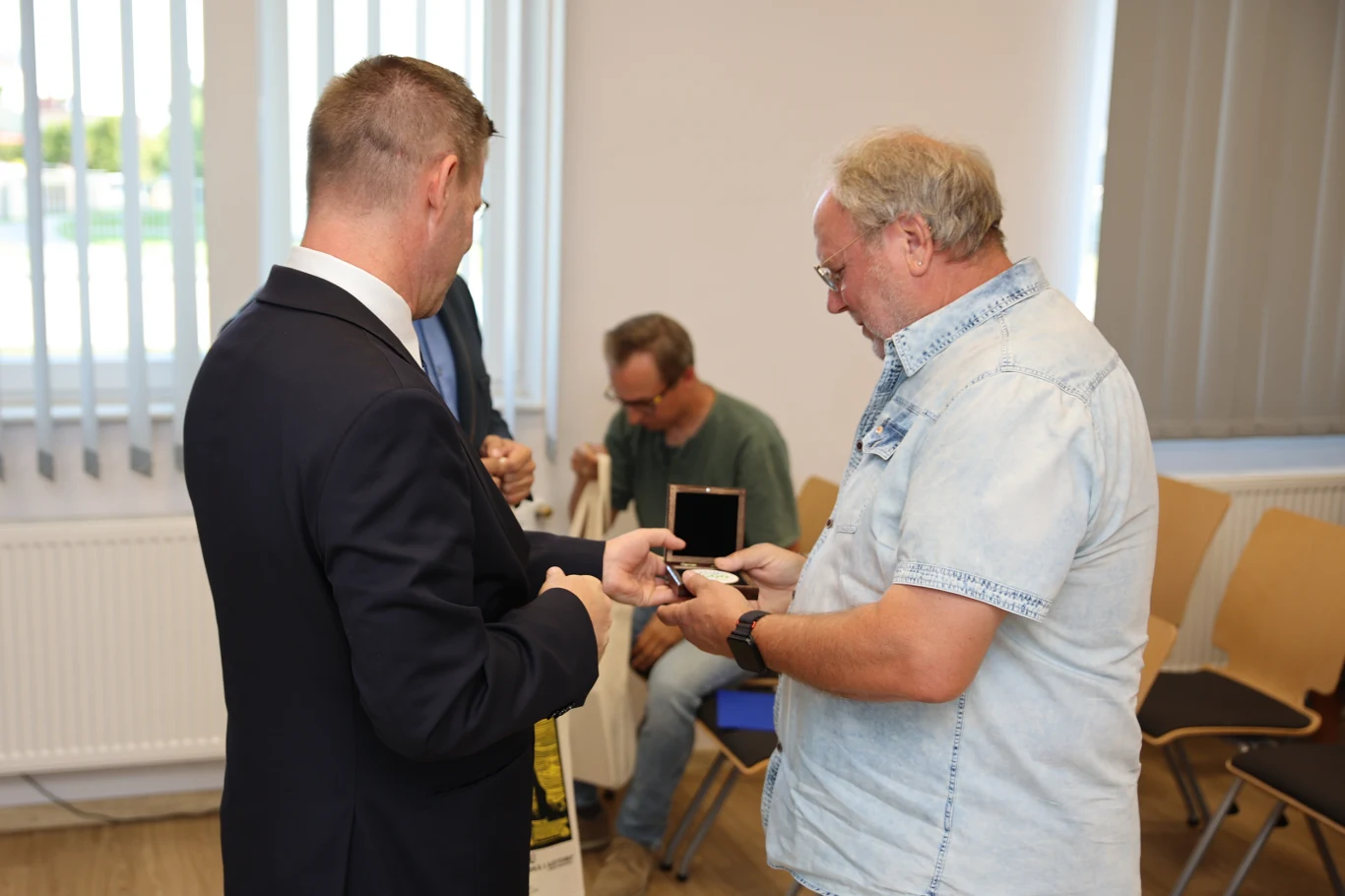 Prezydent Miasta Ełku Tomasz Andrukiewicz odbierający pamiątkę od działacza Solidarności Dwóch mężczyzn przekazujących sobie medal w jasnym, urzędowym pomieszczeniu, w tle siedząca osoba i puste krzesła.