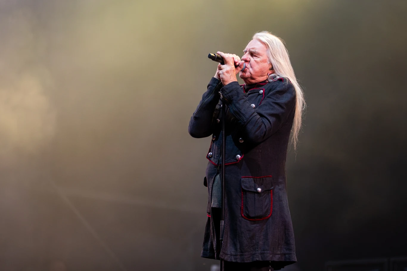 Biff Byford (Saxon) w akcji Biff Byford z Saxon śpiewa do mikrofonu na scenie, długie siwe włosy, czarny płaszcz, kolorowe oświetlenie.