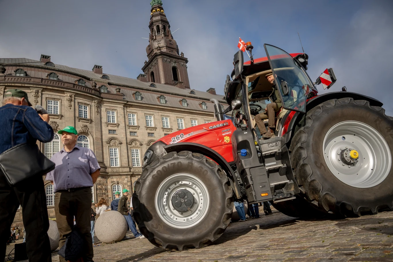 Czerwony ciągnik ustawiony na brukowanym placu przed zabytkowym budynkiem, wokół którego stoją rozmawiający ludzie.