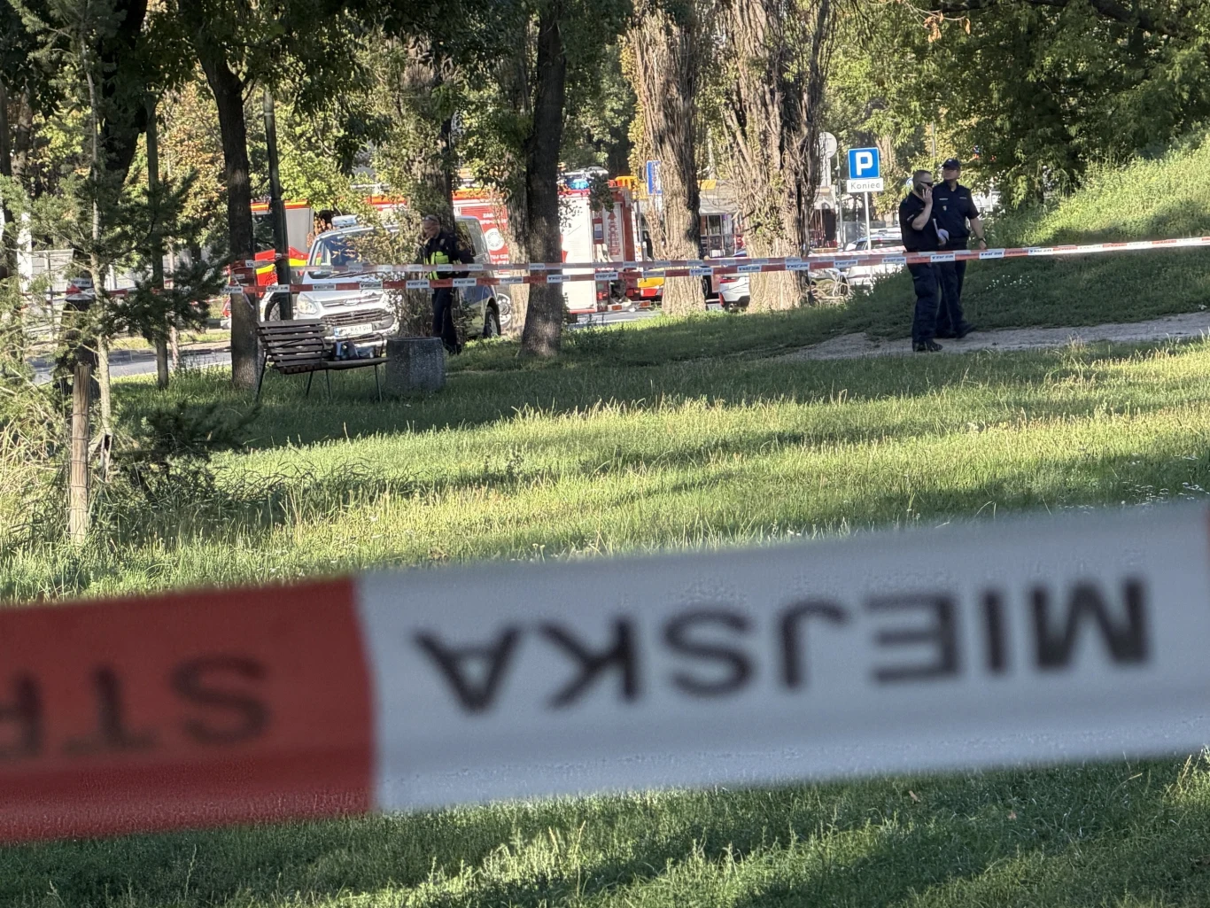 Interwencja służb w związku z odnalezieniem podejrzanych przedmiotów przy ul. Żupniczej w Warszawie Teren zielony odgrodzony taśmą z napisem „Straż Miejska”, w oddali samochody straży pożarnej oraz dwóch funkcjonariuszy stojących przy ścieżce.