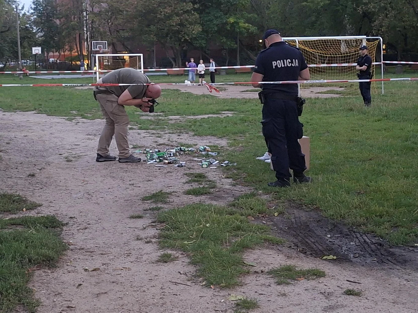 Policyjni technicy badają pakunki przy ul. Żupniczej w Warszawie Policjant oraz fotograf dokumentują rozrzucone na ziemi puszki i butelki w miejscu odgrodzonym taśmą na boisku sportowym, w tle bramki piłkarskie oraz osoby obserwujące sytuację.