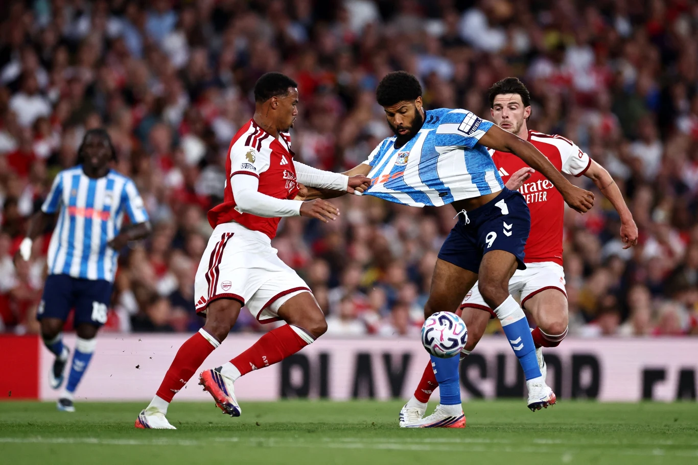 Ellis Simms (nr 9) jako zawodnik Coventry w meczu z londyńskim Arsenalem Dwóch zawodników w czerwono-białych strojach próbuje odebrać piłkę napastnikowi drużyny w niebiesko-białych pasach podczas dynamicznej akcji na murawie stadionu.