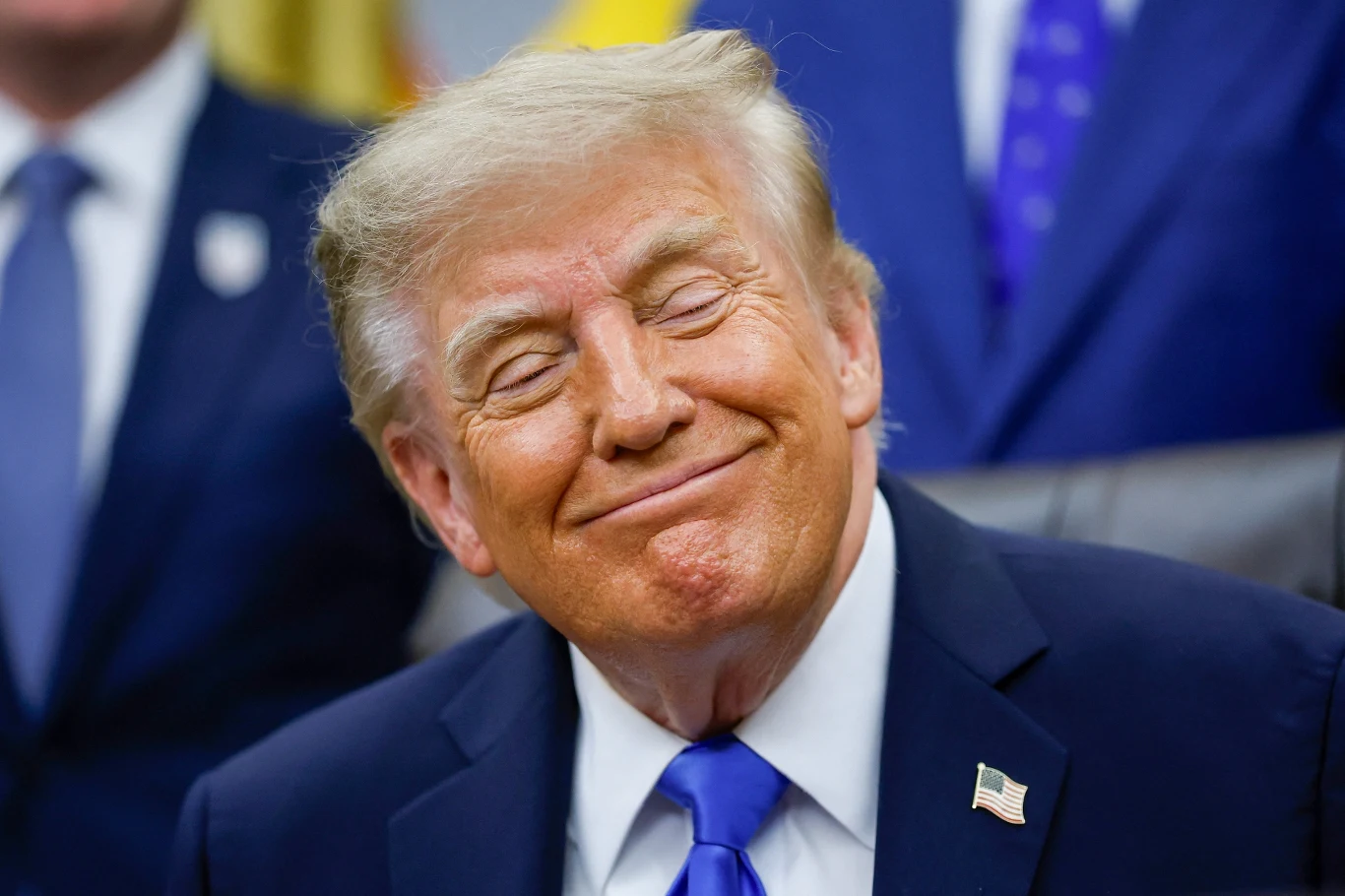 Do obiegu trafiły jednodolarówki z wizerunkiem Donalda Trumpa. Zdj. ilustracyjne Uśmiechnięty Donald Trump w garniturze z flagą USA w klapie siedzi przy biurku.