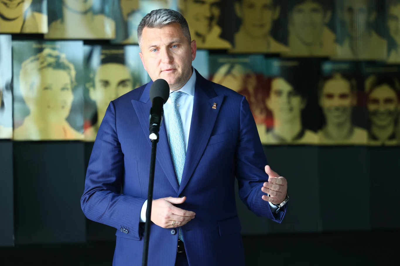 Radosław Piesiewicz Radosław Piesiewicz w granatowym garniturze przemawia do mikrofonu, w tle portrety na żółto-niebieskich planszach