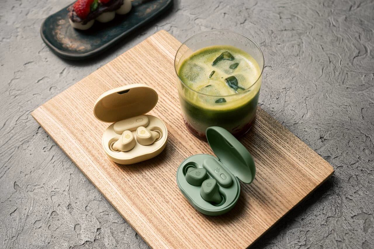SteelSeries x KontrolFreek Słuchawki SteelSeries Arctis GameBuds Matcha i Yuzu w etui na desce, obok szklanki z napojem i miętą.