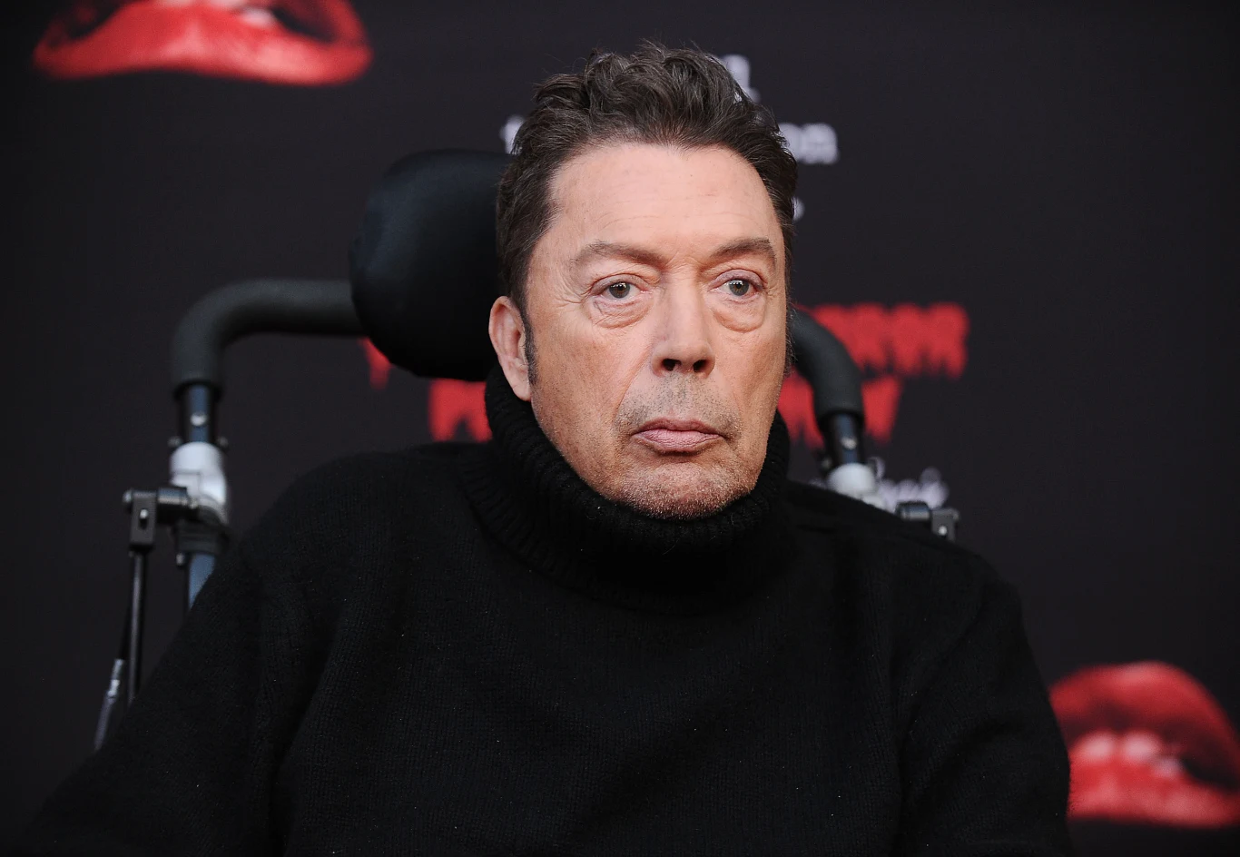 Tim Curry Tim Curry w czarnym golfie na wózku inwalidzkim, na tle czarnej planszy z czerwonymi ustami.