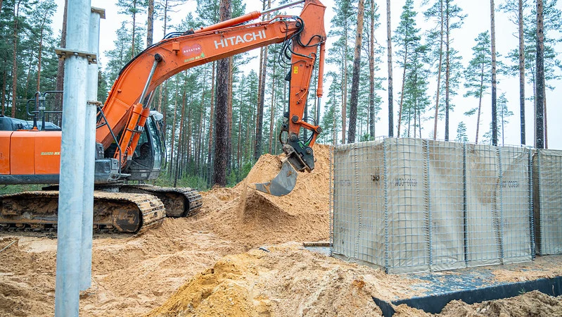 Budowa Bałtyckiej Linii Obrony w Estonii, na granicy z Rosją Koparka gąsienicowa w trakcie prac ziemnych obok stalowego szalunku fundamentowego, w tle widoczne drzewa sosnowe.