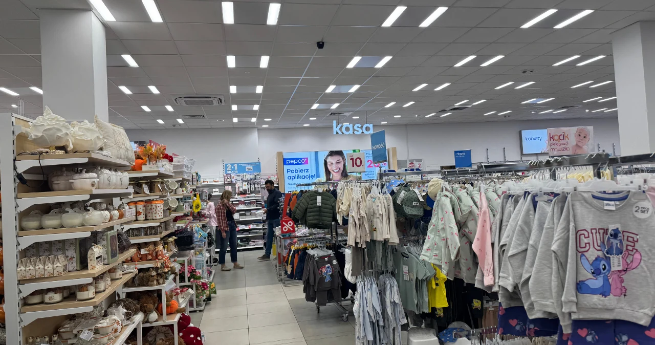 Pepco pokazało nową kolekcję do wnętrz. Kwiatowa elegancja już od 12 zł Regały z ubraniami dziecięcymi po prawej, po lewej dekoracje do domu, w tle kasa i osoby robiące zakupy w Pepco.
