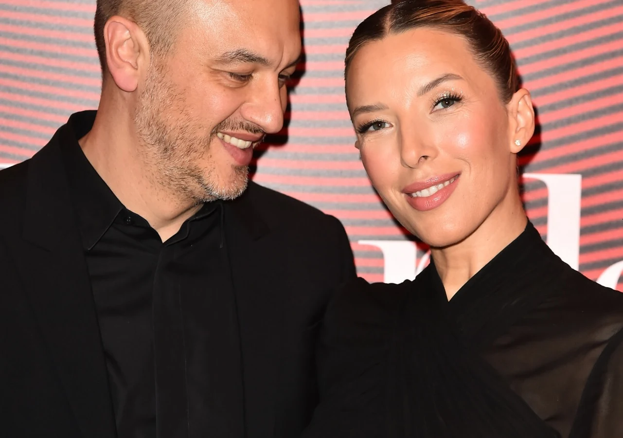 Lefteris Kavoukis i Ewa Chodakowska Uśmiechnięta para ubrana w eleganckie, czarne stroje pozuje razem na tle czerwonej ściany z jasnymi napisami.