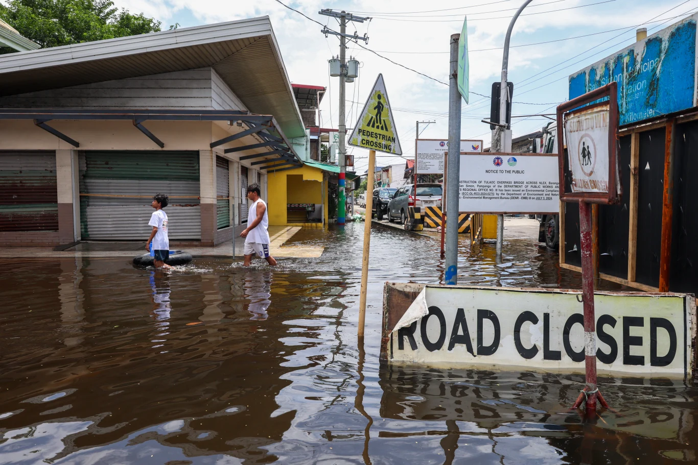 El Nino może przynieść także niszczycielskie powodzie Dwie osoby przechodzą przez zalaną ulicę, rozlewisko przykrywa całą jezdnię, a na pierwszym planie stoi znak informujący o zamknięciu drogi z powodu powodzi.