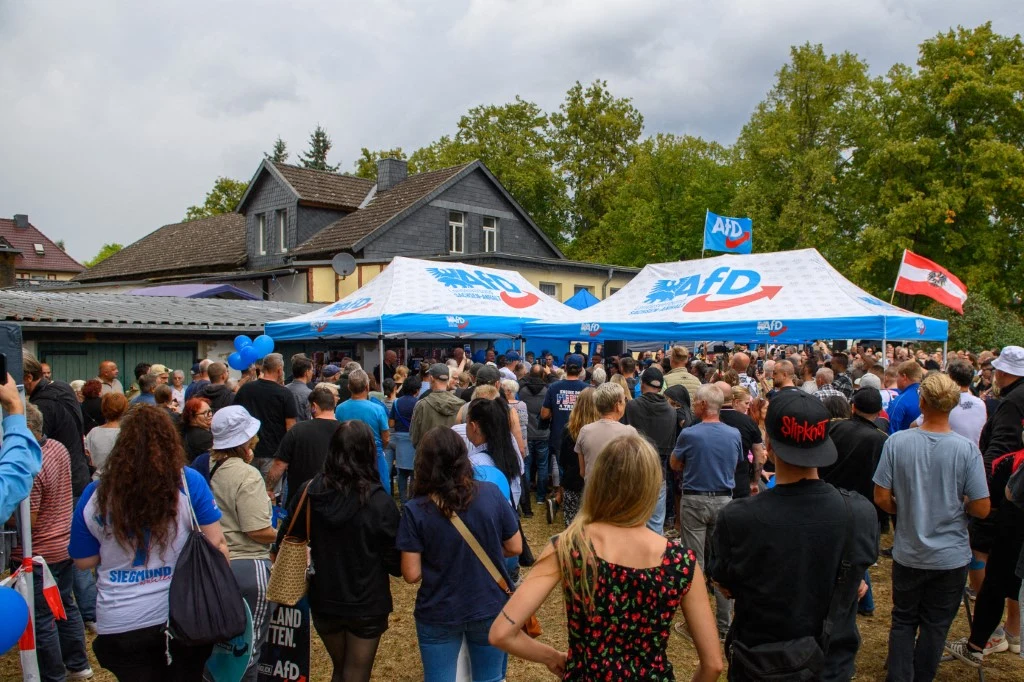 Piknik AfD w Haldensleben Tłum ludzi zgromadzony wokół namiotów z logo partii AfD, w tle widoczne flagi i dom mieszkalny otoczony drzewami.