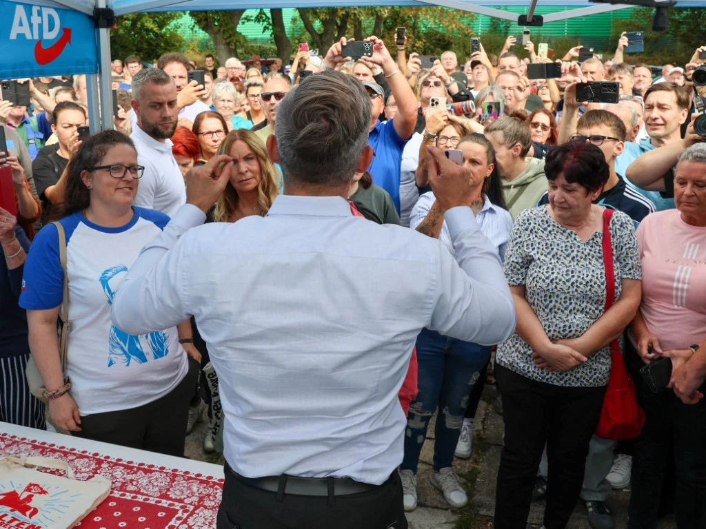 Mężczyzna w białej koszuli przemawia do zgromadzonego tłumu ludzi stojących na zewnątrz, część osób robi zdjęcia lub nagrywa telefonami, w tle widoczny namiot z logotypem AfD.