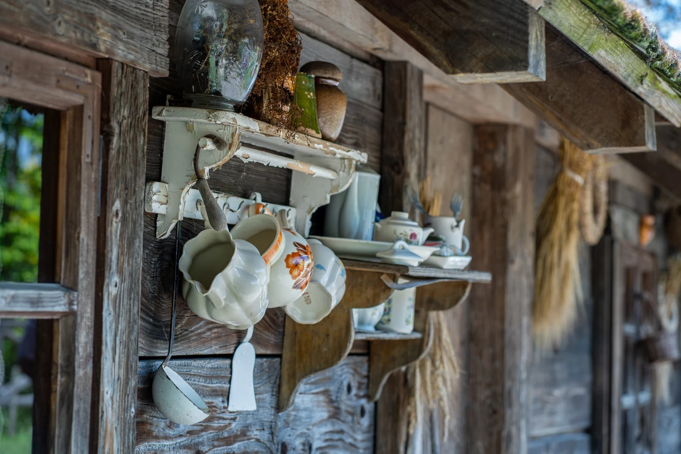 Zabytkowa półka z porcelanowymi dzbankami i filiżankami zawieszona na drewnianej ścianie wiejskiej chaty, obok wiszących kłosów zbóż.