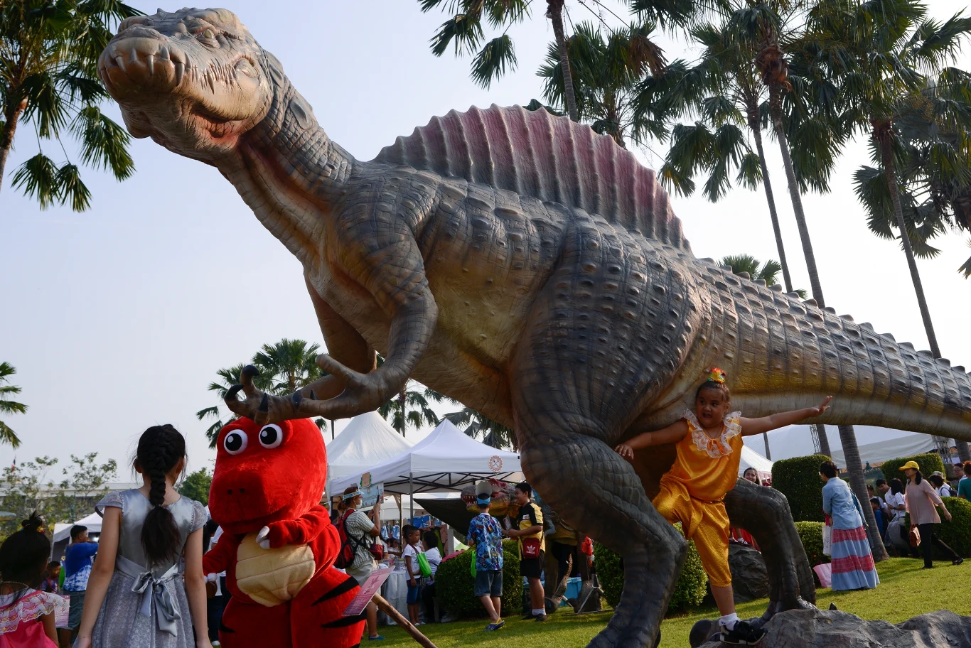 Duża figura dinozaura otoczona przez dzieci, z których jedno stoi w przebraniu czerwonego smoka, a inne pozują obok monumentu na terenie parku wśród palm.