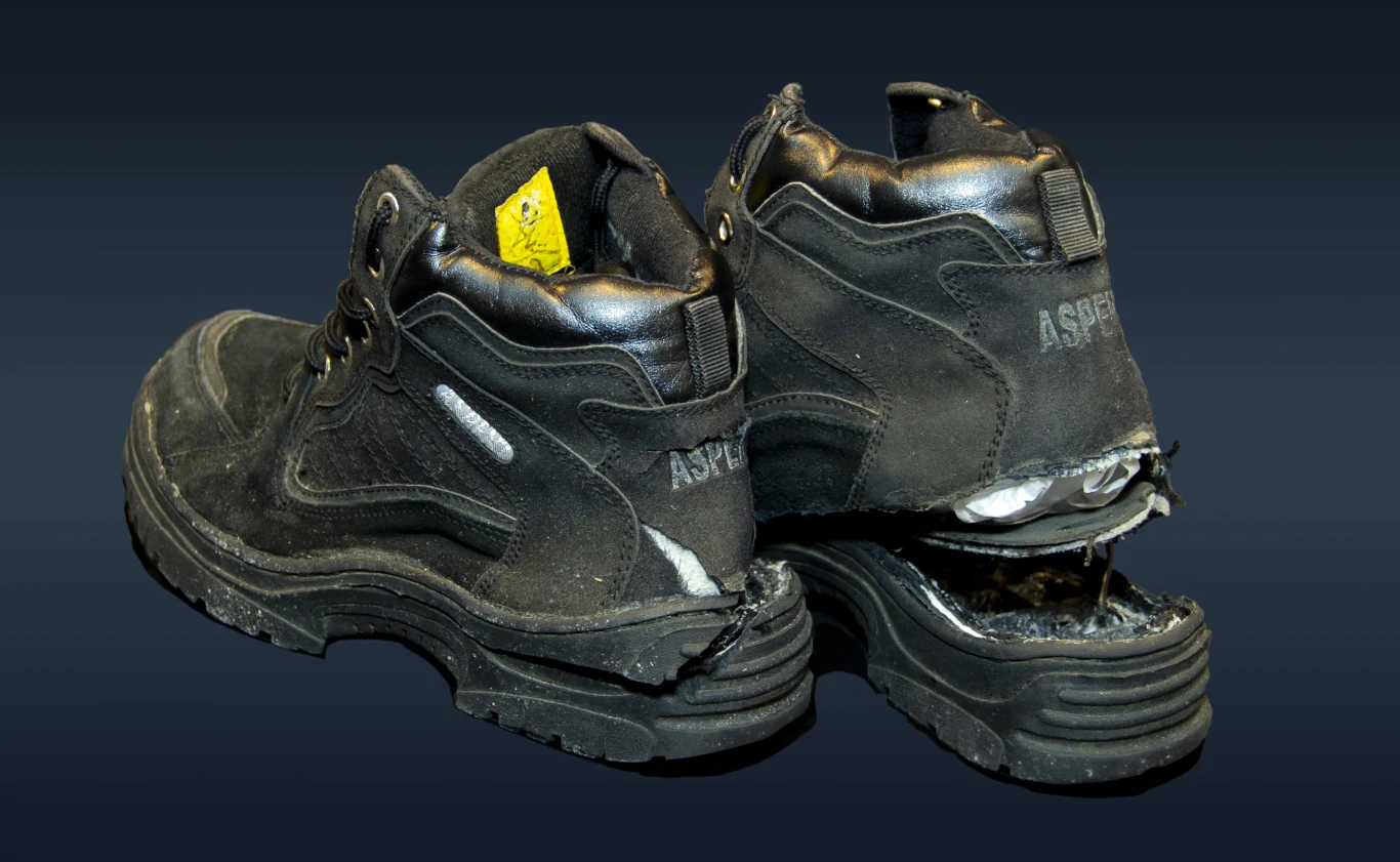 Buty, które Reid próbował zdetonować w czasie lotu. Czarne, mocno zużyte robocze buty z wyraźnymi uszkodzeniami w tylnej części jednej z podeszw, z widocznym elementem żółtej metki wewnątrz jednego buta.