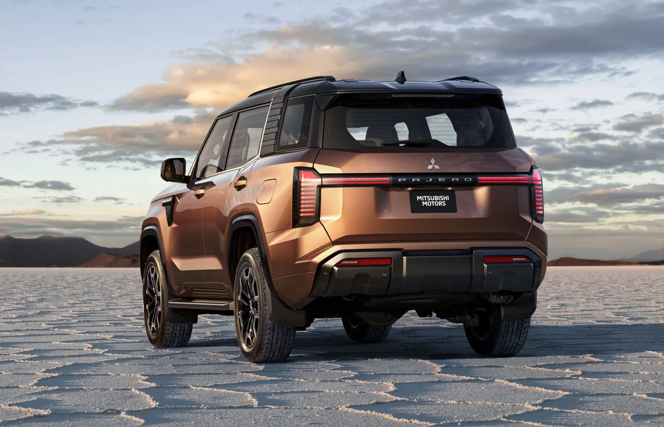 Brązowy SUV Mitsubishi stoi na płaskiej, pustynnej powierzchni z wyraźnymi heksagonalnymi formacjami, w tle niskie góry i niebo z rozproszonymi chmurami.