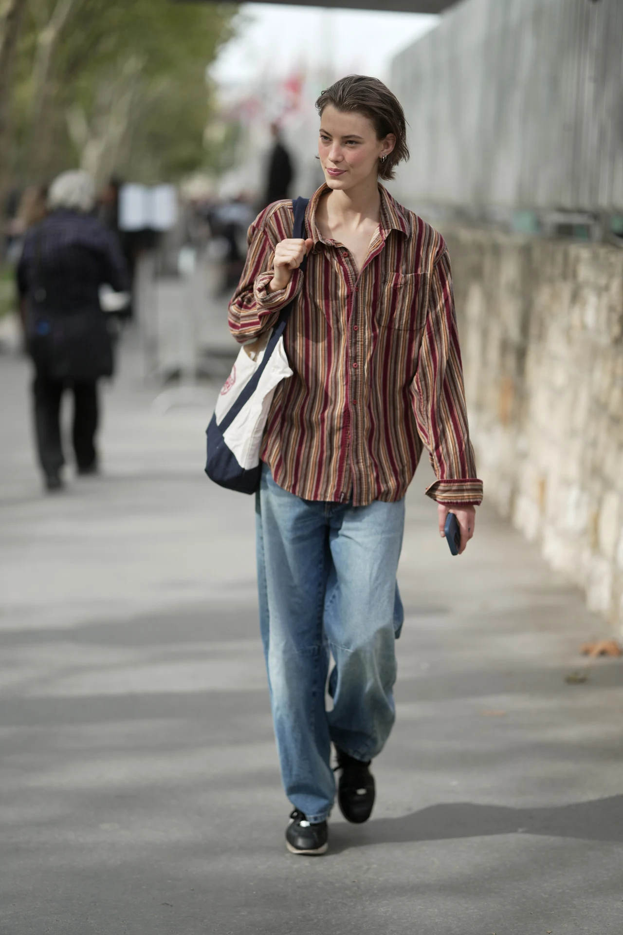Dana Smith uzupełniła stylizację torbą Trader Joe's na Paris Fashion Week 2025 Młoda osoba w casualowej, prążkowanej koszuli i szerokich jeansach idzie chodnikiem, trzymając w ręku telefon i przewieszoną przez ramię torbę na zakupy.