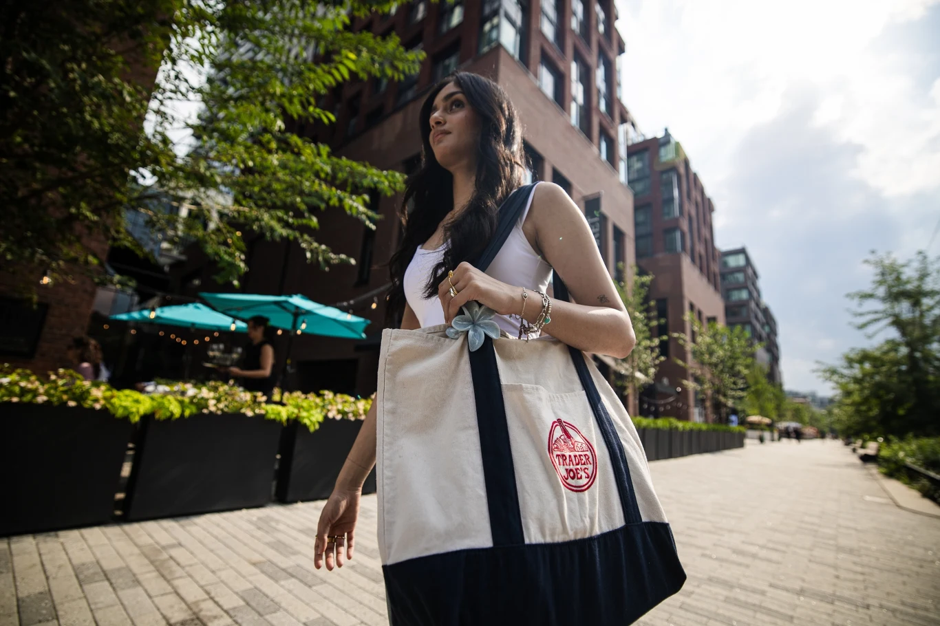 Biedronka wypuściła własną torbę inspirowaną shopperką Trader Joe's Kobieta z długimi ciemnymi włosami spacerująca po miejskiej alejce, trzyma duży materiałowy worek z logo Trader Joe’s; w tle nowoczesne budynki i zielone rośliny na tarasie restauracji.