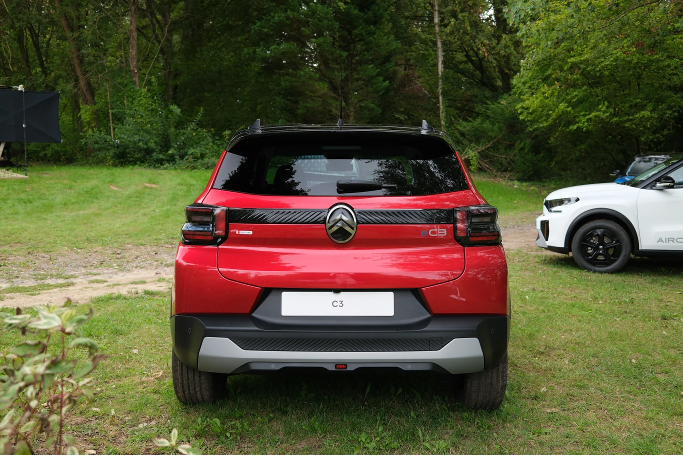 Czerwony Citroën C3 widziany od tyłu, ustawiony na trawie w pobliżu białego samochodu, otoczony zielenią drzew i krzewów.
