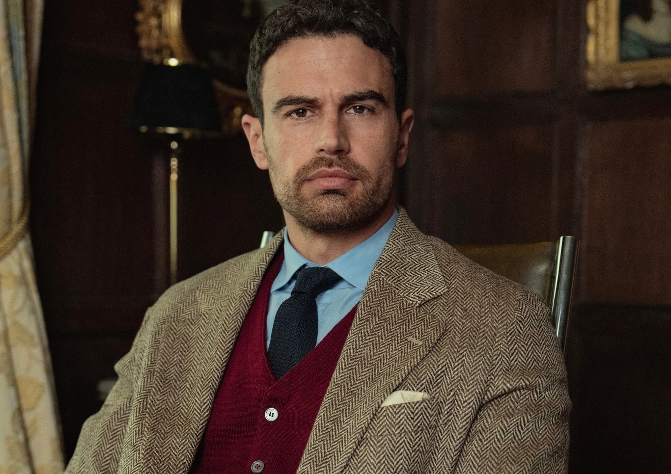 "Dżentelmeni": Theo James jako Eddie Theo James jako Eddie siedzi na krześle w brązowej marynarce, bordowym swetrze i krawacie w eleganckim wnętrzu.