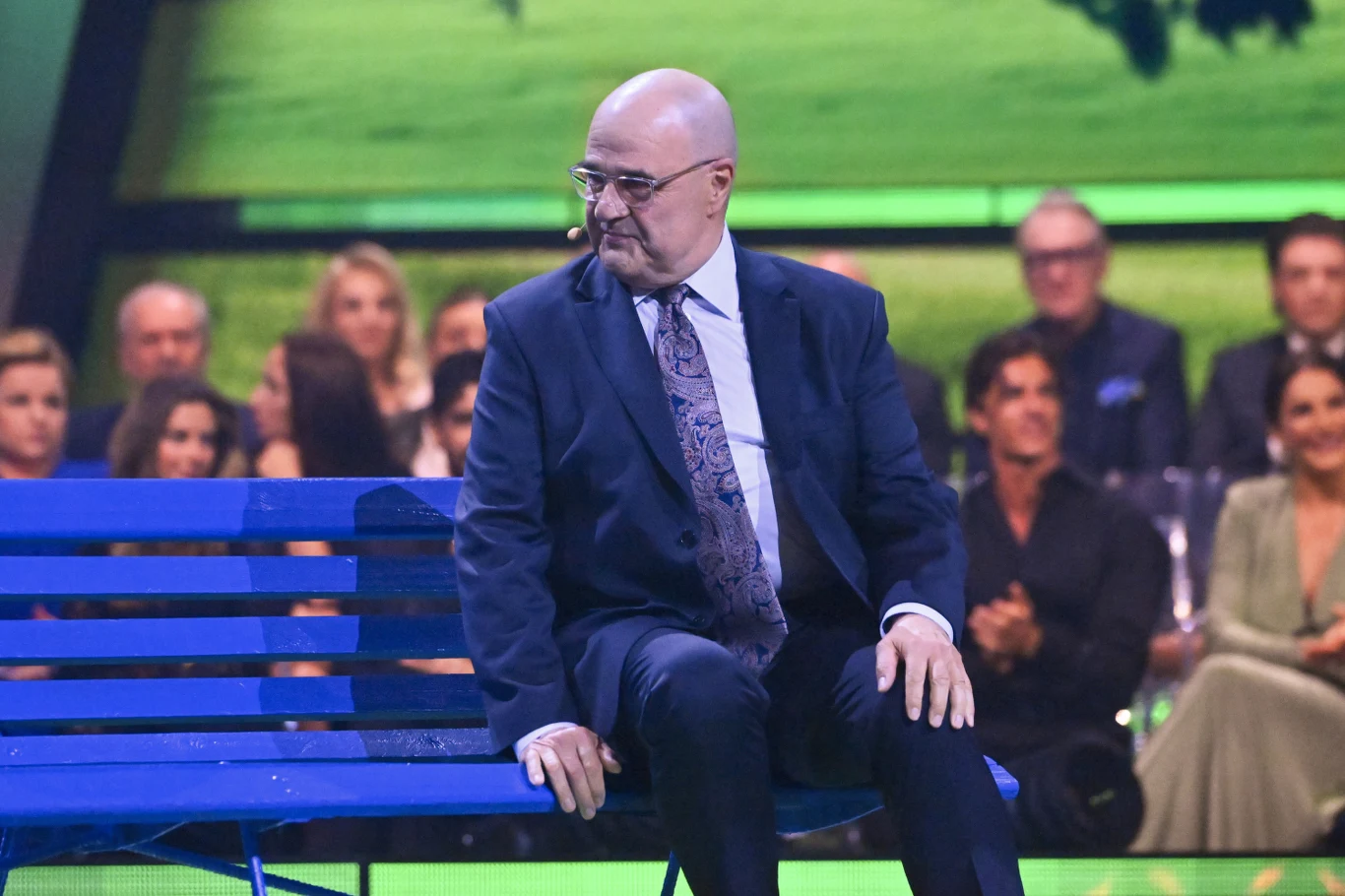 Cezary Żak na ramówce TVP Łysy mężczyzna w garniturze i z mikrofonem przy ustach siedzi na niebieskiej ławce, a w tle widać publiczność w eleganckich strojach.