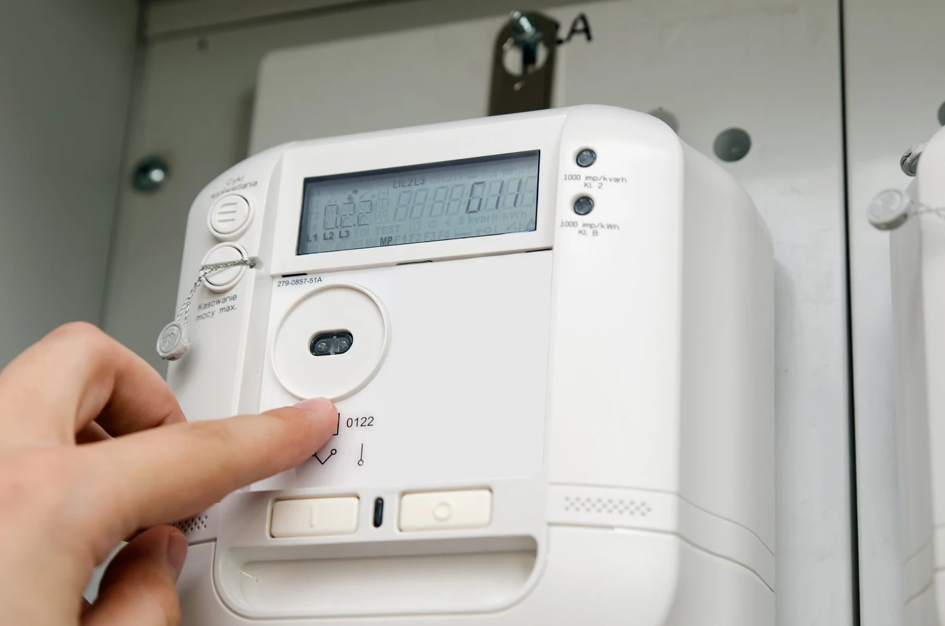 Jak odczytywać liczby na liczniku prądu? Elektroniczny licznik prądu z wyświetlaczem i przyciskami, ręka zbliżająca palec do urządzenia