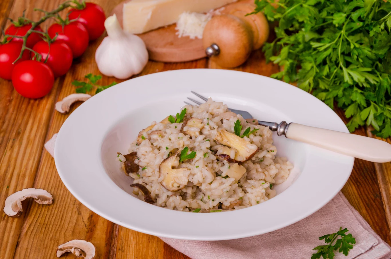 To prawdziwa klasyka kuchni włoskiej Risotto z grzybami, natką pietruszki, podane w głębokim talerzu na stole; obok pomidory, czosnek, parmezan, pieczarki.