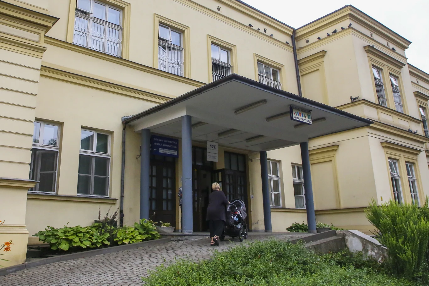 Wejście do dużego budynku z charakterystycznym daszkiem wspartym na dwóch słupach, kobieta z wózkiem dziecięcym kieruje się do głównych drzwi.