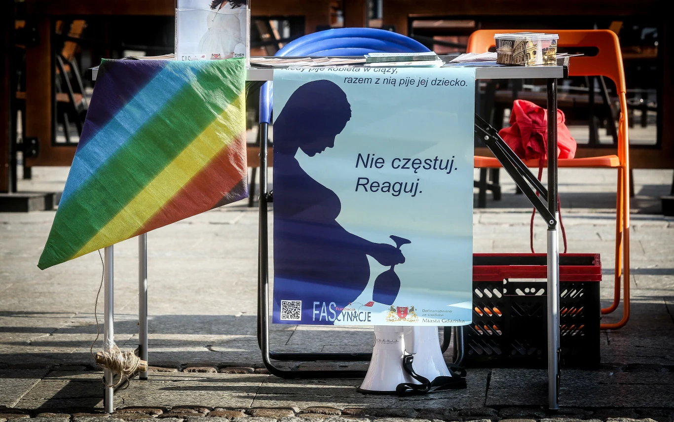 Stolik uliczny z przyczepioną tęczową flagą oraz dużym plakatem kampanii informacyjnej, na którym widnieje sylwetka ciężarnej kobiety trzymającej butelkę, opatrzona napisem: „Nie częstuj. Reaguj.”