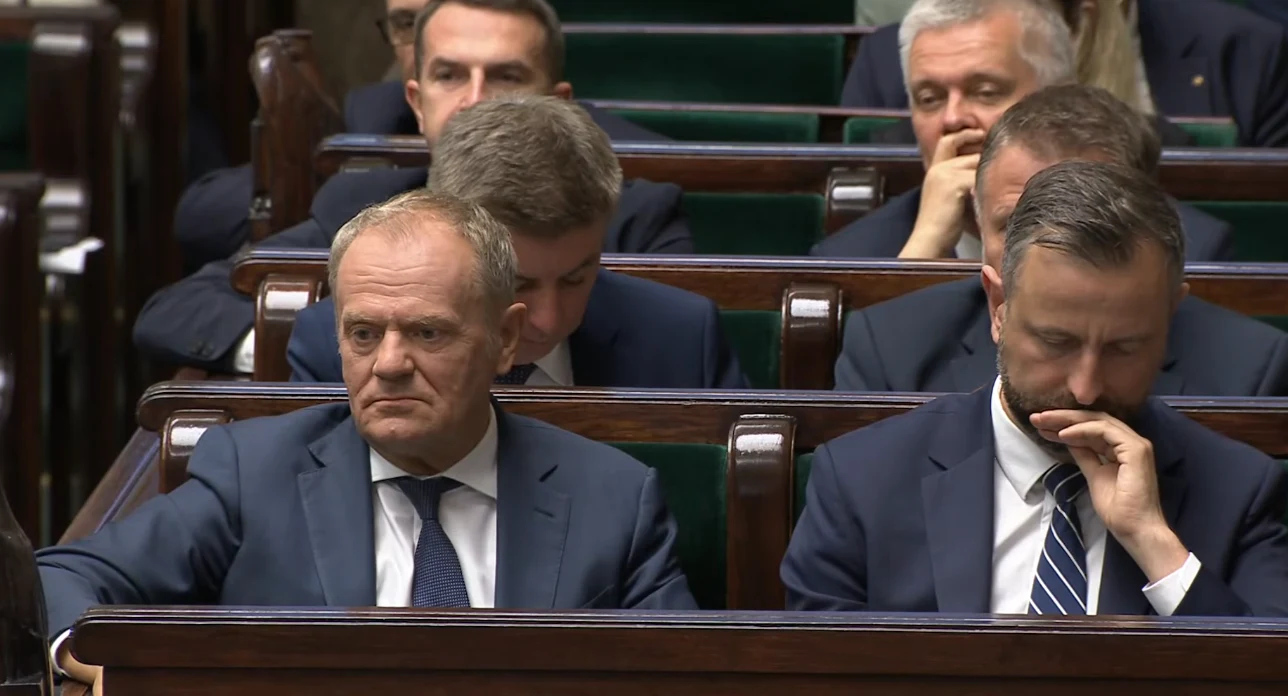 Grupa mężczyzn ubranych w garnitury siedzi w ławach sejmowych, skoncentrowani i zamyśleni, z poważnymi wyrazami twarzy.